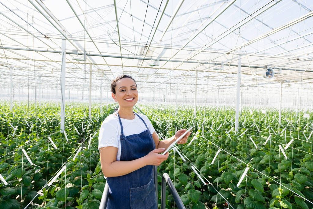 happy african american farmer in apron standing with digital tablet in glasshouse