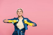 Positive Frau in Gummihandschuhen hält Waschmittel und Lappen isoliert auf rosa