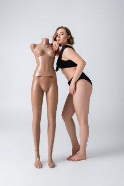 full length of overweight and barefoot woman in swimsuit leaning on plastic mannequin on white  clipart
