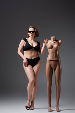 full length of overweight young woman in swimsuit and sunglasses posing near plastic mannequin on grey clipart