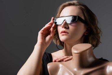 overweight young woman adjusting sunglasses and posing with plastic mannequin isolated on grey clipart