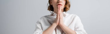 cropped view of young overweight woman in white shirt with praying hands isolated on grey, banner clipart