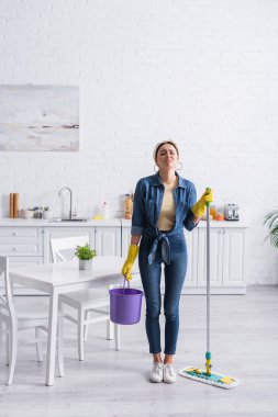 Displeased housewife holding bucket and mop in kitchen  clipart