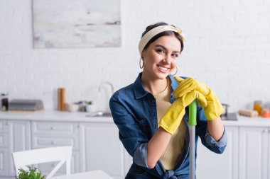 Housewife in rubber gloves holding mop and smiling at camera  clipart