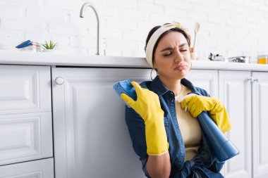 Displeased housewife in rubber gloves holding sponge and detergent in kitchen  clipart