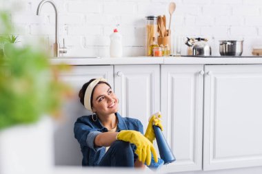 Lastik eldivenli neşeli kadın mutfakta bir şişe deterjan ve sünger tutuyor. 
