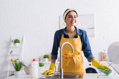 Gülümseyen kadın tabağın yanında bez tutuyor ve mutfakta lavabo açıyor. 