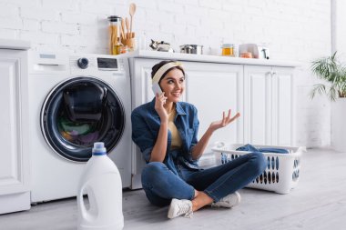 Smiling housewife talking on smartphone near detergent and washing machine in kitchen  clipart