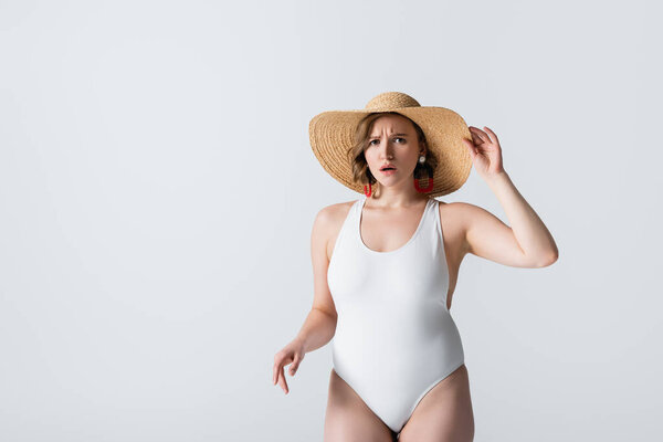 толстая и шокированная женщина в сережках и купальнике, регулируя соломенную шляпу, изолированную на белом