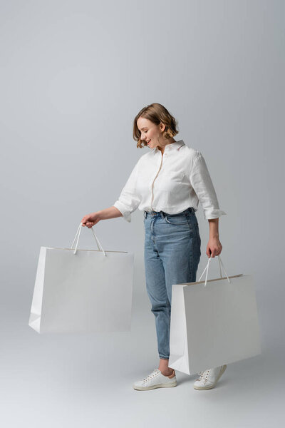 happy overweight woman in jeans and white shirt holding shopping bags on grey 