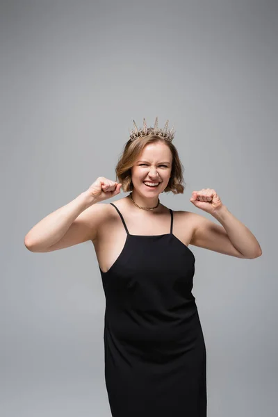 happy plus size woman in black slip dress and crown isolated on grey