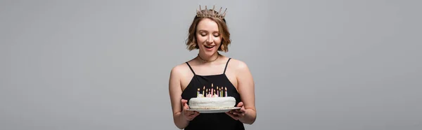 happy plus size woman in slip dress and crown holding birthday cake isolated on grey, banner