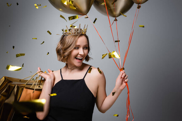 happy plus size woman in slip dress and crown holding balloons and shopping bags near confetti on grey 