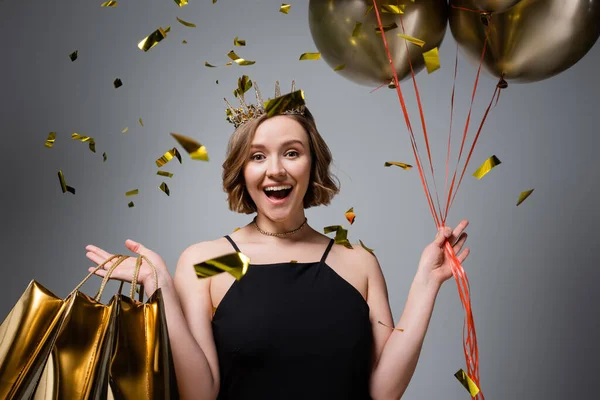 amazed plus size woman in slip dress and crown holding balloons and shopping bags near confetti on grey