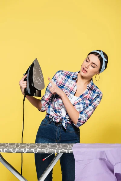 Housewife pointing with finger at iron near board on yellow background