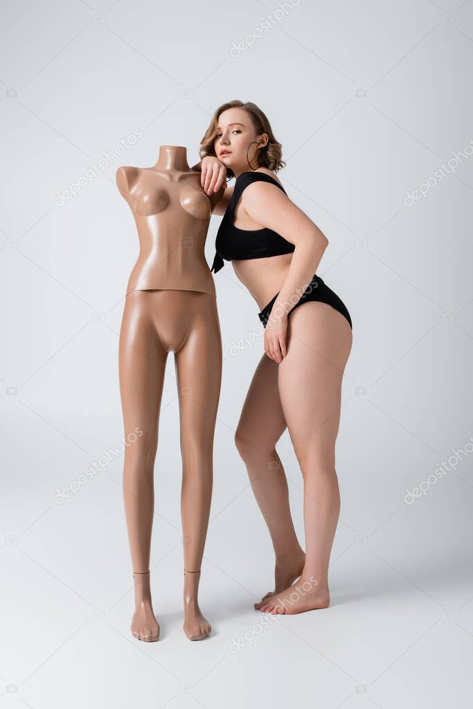 full length of overweight and barefoot woman in swimsuit leaning on plastic mannequin on white 