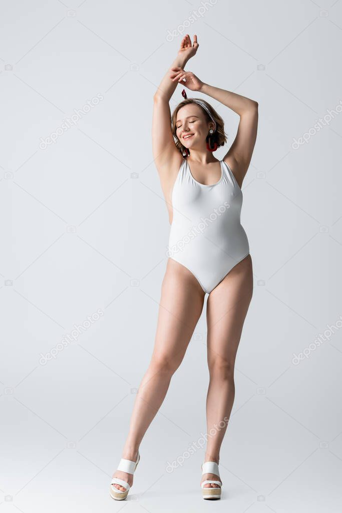 full length of smiling overweight young woman in swimwear and earrings posing on white