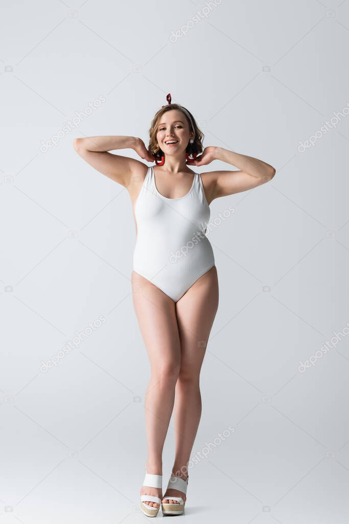 full length of cheerful overweight young woman in swimwear and earrings smiling while posing on white