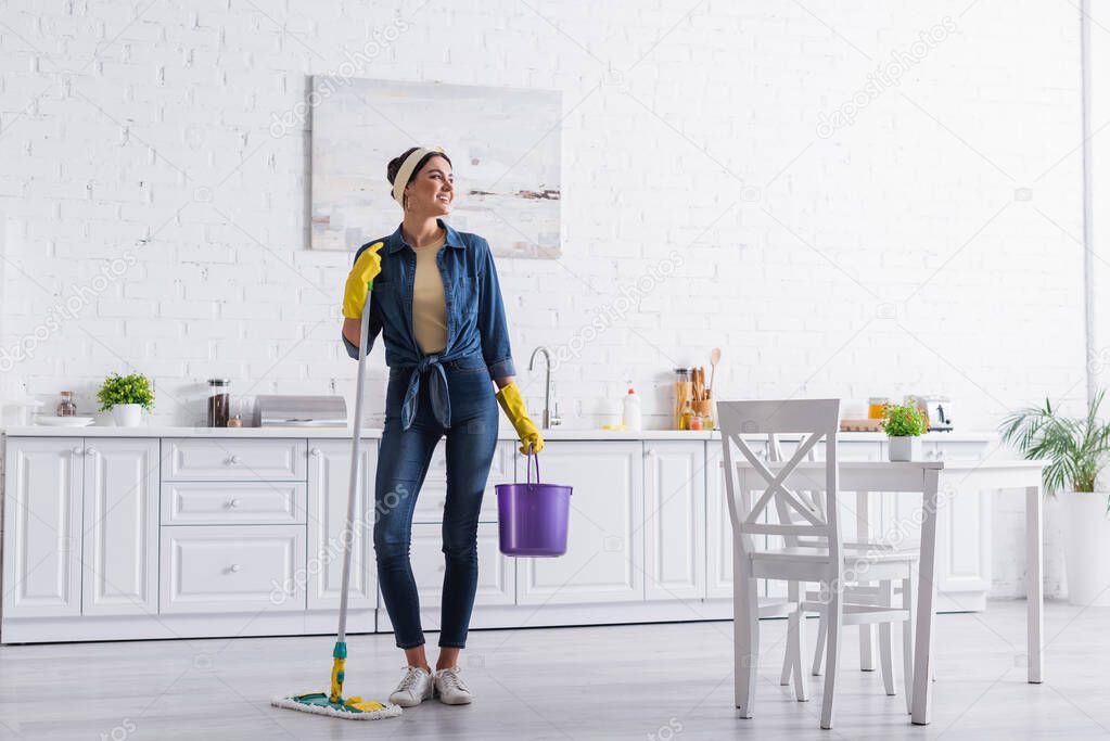 Positive housewife holding bucket and mop in kitchen 