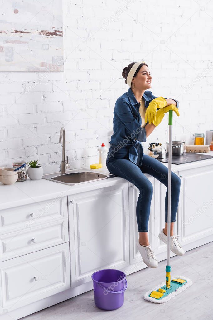 Smiling woman in rubber gloves holding mop while sitting on kitchen worktop 