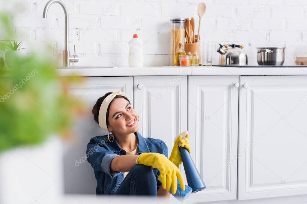 Cheerful woman in rubber gloves holding bottle of detergent and sponge in kitchen 