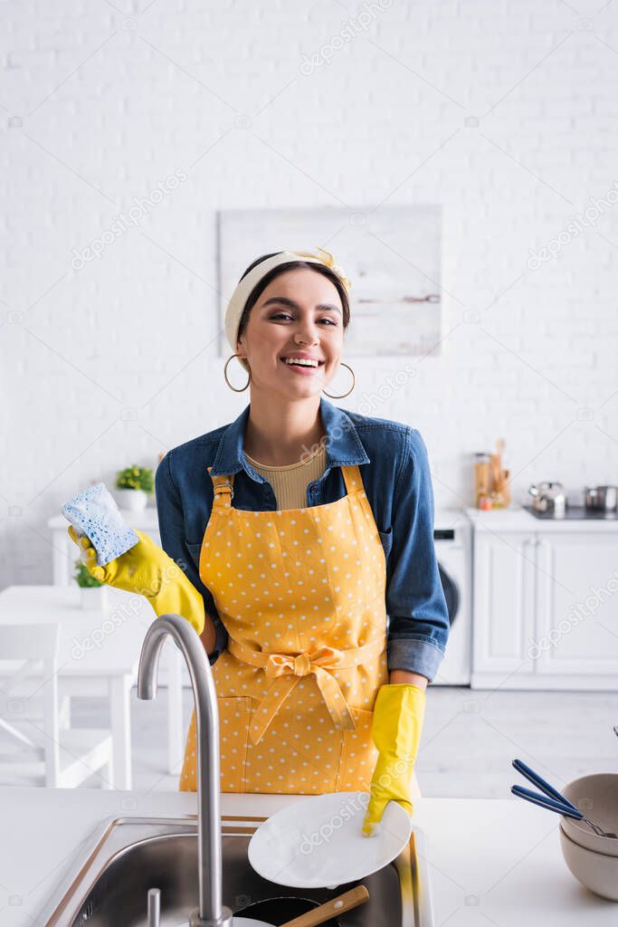 Cheerful housewife with sponge and plate looking at camera near sink 