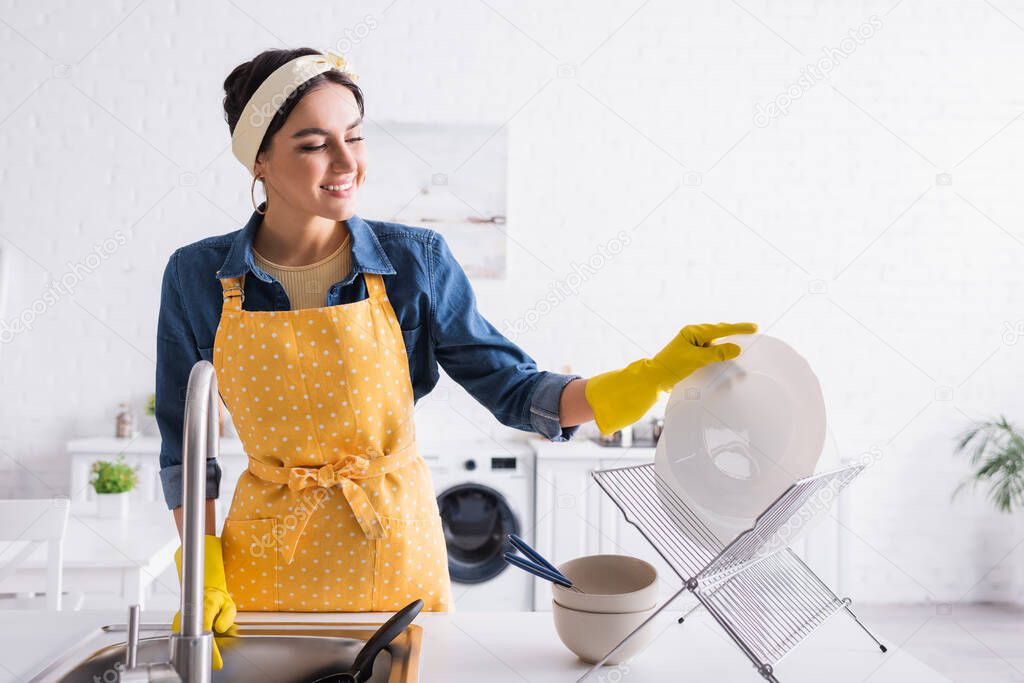 Happy woman in apron putting plate on stand in kitchen 