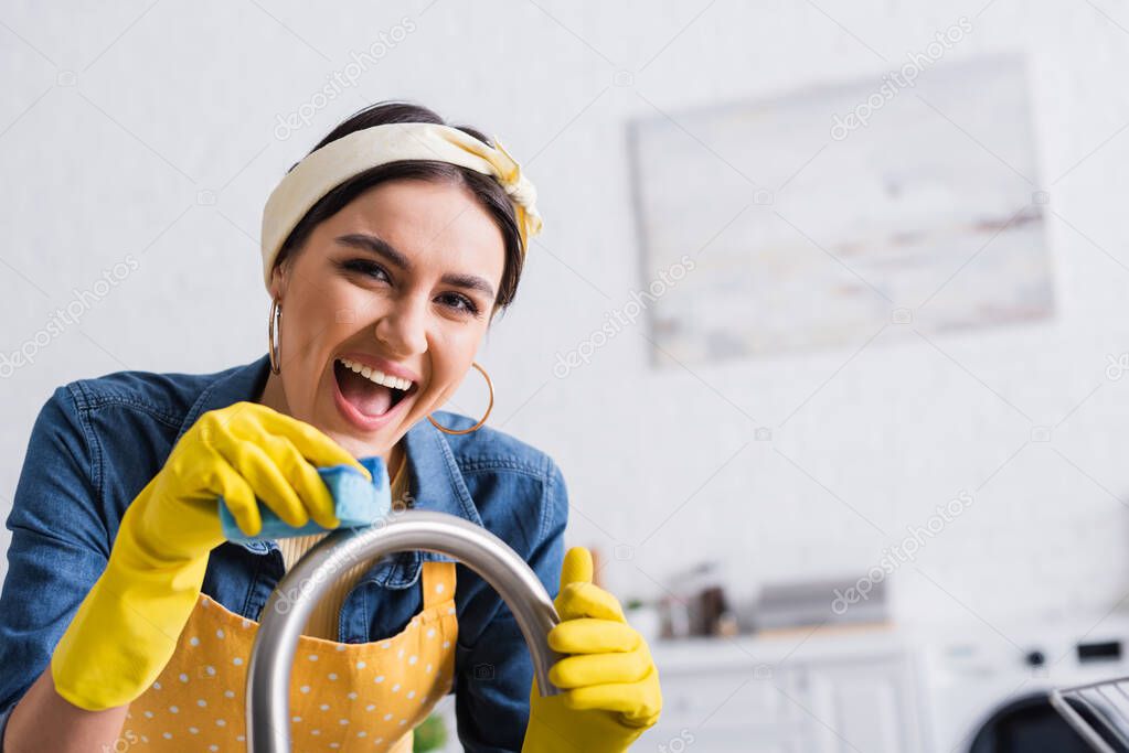 Cheerful woman cleaning kitchen faucet with sponge 