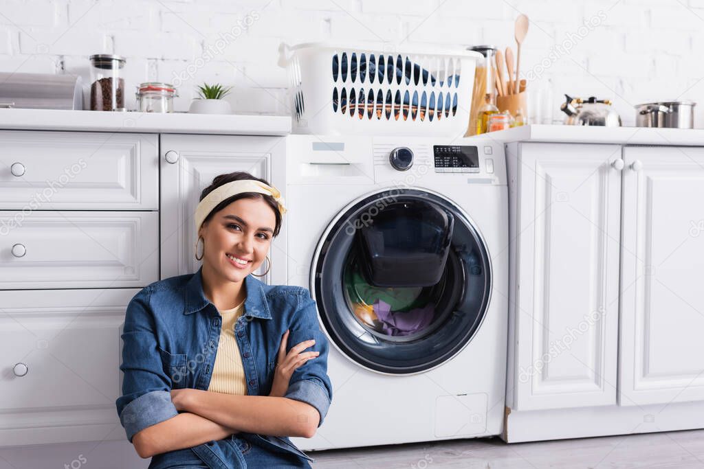 Smiling housewife looking at camera near washing machine in kitchen 