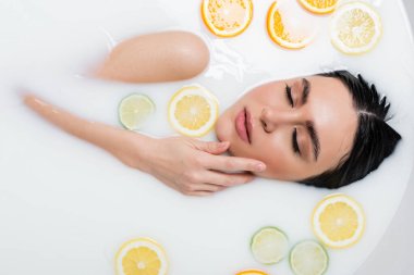 top view of young woman touching face while bathing in milk with citrus slices clipart