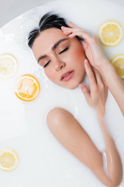 Kapalı gözlü genç bir kadın yüzüne yakın el ele tutuşurken limon dilimleriyle süt banyosunun tadını çıkarıyor.