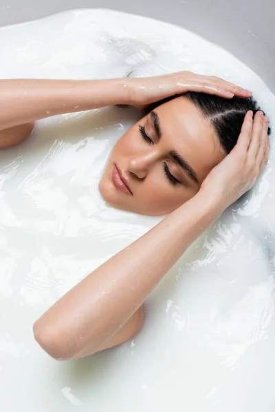 young woman with closed eyes relaxing in milk bath and touching wet hair