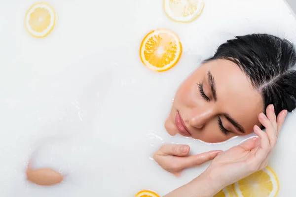 top view of pretty woman with closed eyes taking milk bath with lemon and orange slices