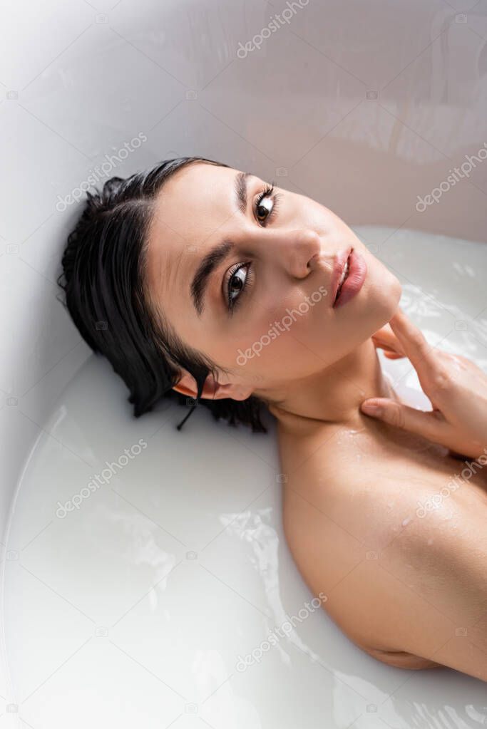 young, sensual woman touching neck and looking at camera in milky bath