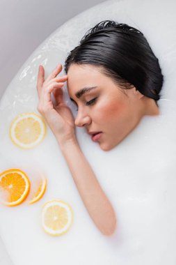 Şehvetli bir kadın süt banyosunda portakal ve limon dilimleriyle gözleri kapalı dinleniyor.