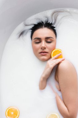 Kapalı gözlü genç bir kadın süt banyosunda dinlenirken portakal dilimini tutuyor.