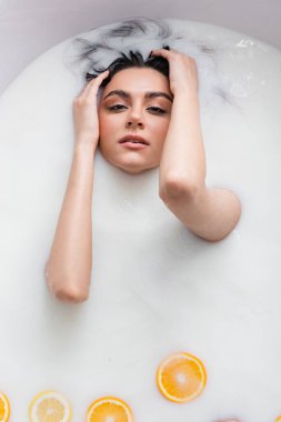 Güzel bir kadın süt banyosunda narenciye dilimleriyle dinlenirken kameraya bakıyor.