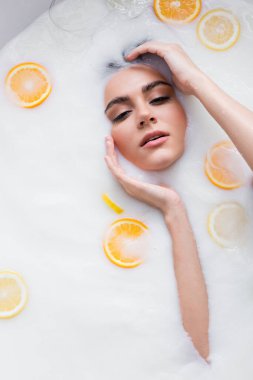 Elleri yüzüne yakın genç bir kadın süt banyosunda dilimlenmiş narenciye meyveleriyle dinleniyor.