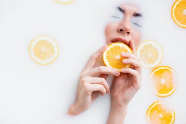high angle view of woman holding orange slice while immersing face in milk bath clipart