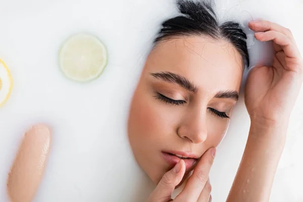 young woman touching face while relaxing in milk bath with citrus slices