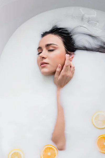 young woman with closed eyes touching face while bathing in milk with sliced citruses