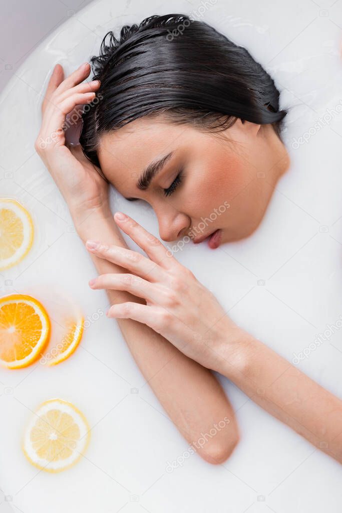 young woman with closed eyes relaxing in milk bath with sliced orange and lemon