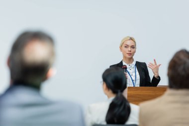 young speaker gesturing while talking to blurred business people during seminar clipart
