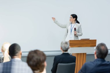 Seminer sırasında Asyalı konuşmacı mikrofonu tutuyor ve eli ile bulanık seyircinin yakınına doğrultuyor.