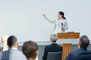 asian lecturer holding microphone and pointing with hand near blurred audience during seminar clipart