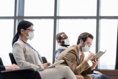 young businessman in medical mask writing in notebook near interracial colleagues clipart