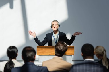 Gülümseyen konuşmacı konferans sırasında açık kollarla bulanık ön plandaki katılımcıların yanında duruyor