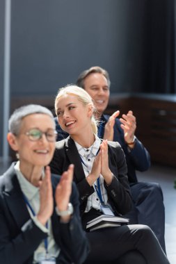 selective focus of young businesswoman smiling and applauding near colleagues clipart