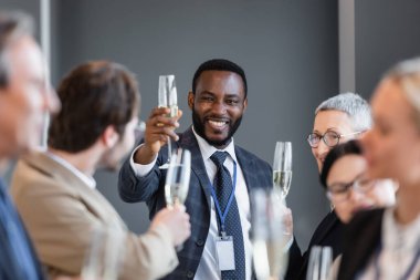 selective focus of african american businessman holding champagne glass near interracial colleagues clipart