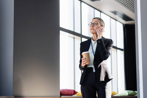 Mature businesswoman holding takeaway drink and talking on smartphone in office 